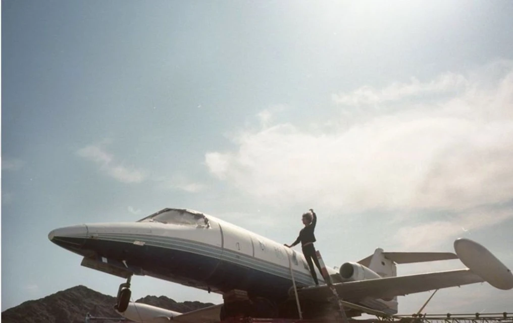 Adult stands atop private jet under a blue sky with white clouds. The person in a dark jumpsuit poses on the wing of the plane.