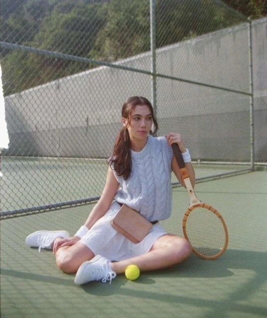 A woman in tennis attire sits on a green tennis court with a racket, wearing a white skirt, light blue vest, and white sneakers outdoors.