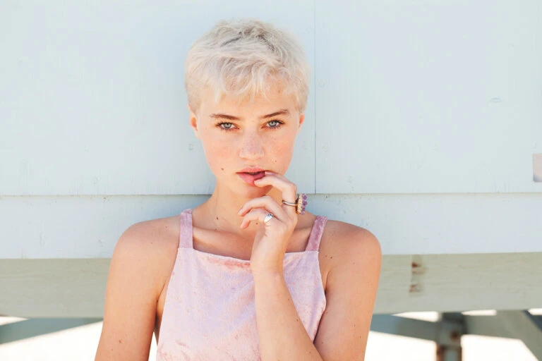 Close-up portrait of an adult woman with short blonde hair and light eyes, wearing a pink dress, posing outdoors against a light-blue wall.