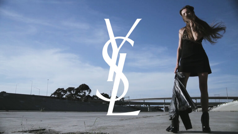 Fashion shot of a young adult woman in a black dress standing on a concrete surface holding a black jacket with the YSL logo in the sky.