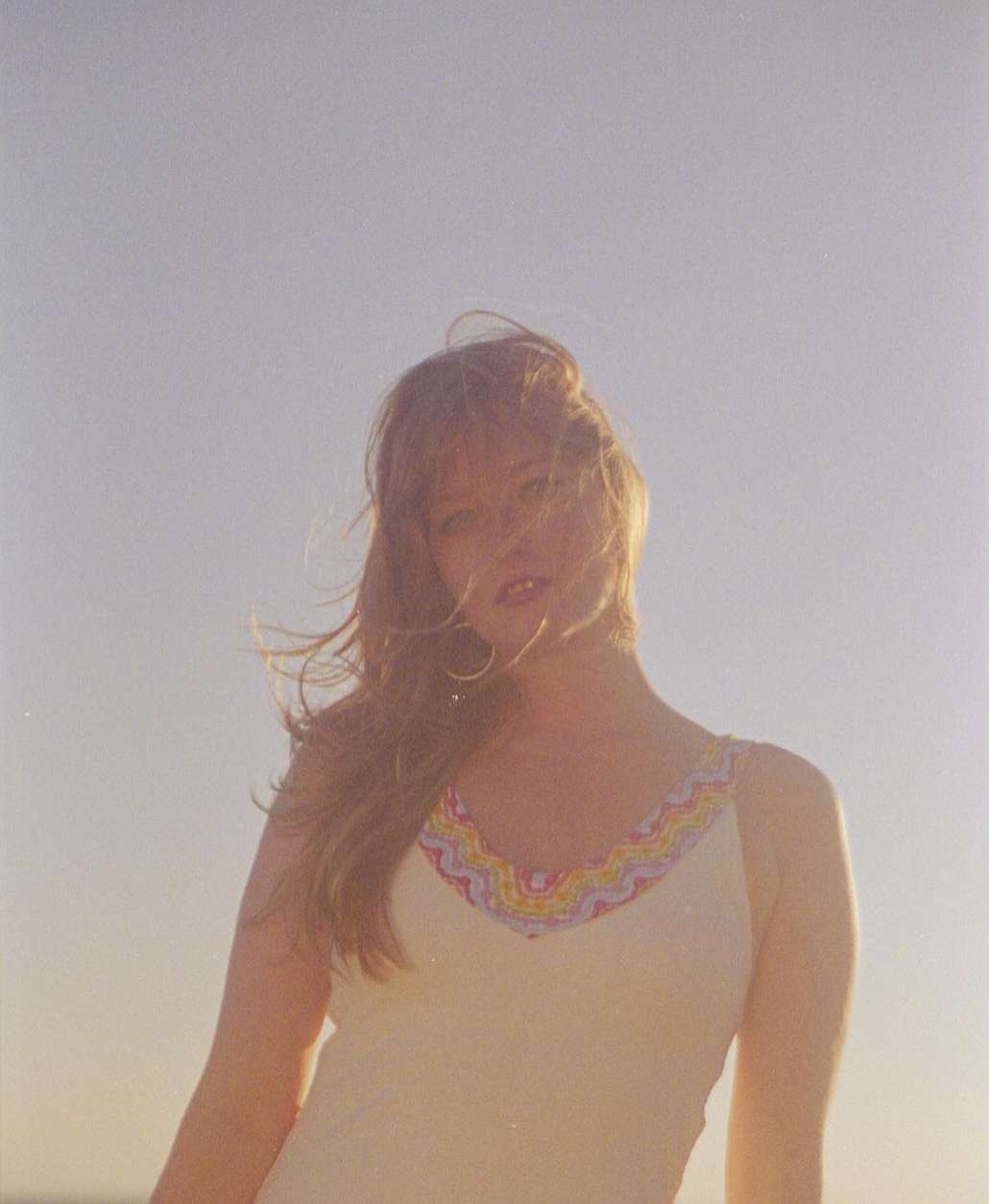 Woman in white tank top gazes toward the camera against a bright sky background, with her hair gently blowing in the wind.