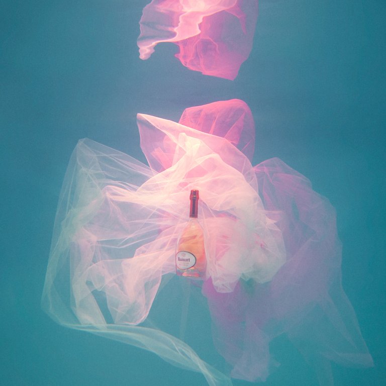 A bottle floats underwater amidst transparent plastic bags, creating a surreal and ethereal scene with soft, diffused lighting and gentle hues.