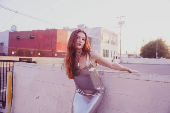 Woman in a glossy metallic silver dress leaning against a low white wall outdoors during sunset by Nastasia Dusapin