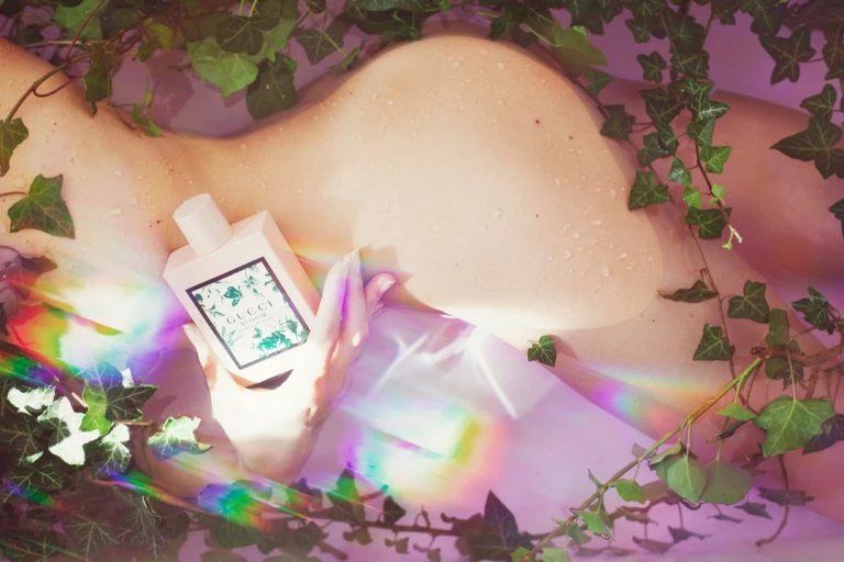 Perfume bottle held by a person amidst green ivy vines and a rainbow light effect, creating a beauty-focused, artistic composition.