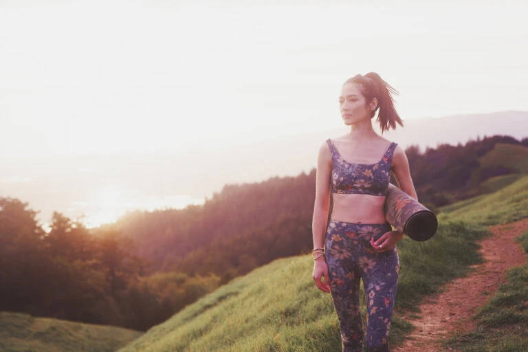 Adult woman in athletic wear walks along a grassy hill at sunset while carrying a rolled-up yoga mat; mountains in the background.