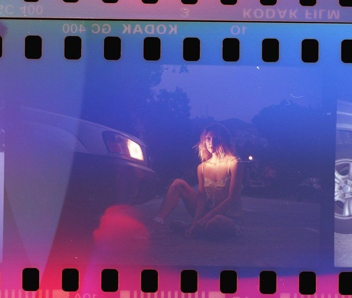 Woman sits near a car at dusk in a photograph showing film borders. The car headlights are on, casting a warm glow.