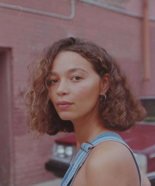 Portrait of a young adult woman with curly hair, wearing denim overalls, standing outdoors with a red brick building in the background.