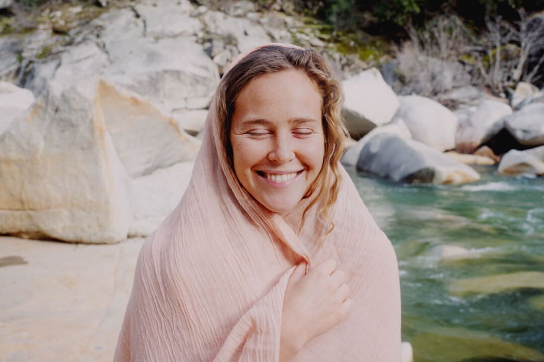 Smiling adult woman with eyes closed wrapped in peach-colored fabric, outdoors near rocks and flowing river during daytime.