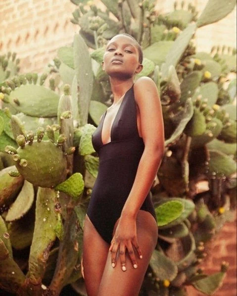 A slender woman in a black swimsuit poses outdoors next to a tall, green cactus. The image is a medium shot with soft, diffused lighting.