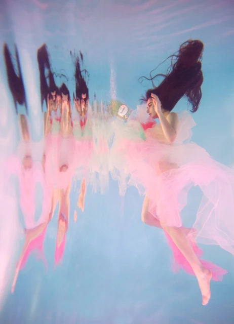 An ethereal underwater portrait captures a woman with flowing dark hair and a light pink dress, floating weightlessly in blue water with soft reflections.