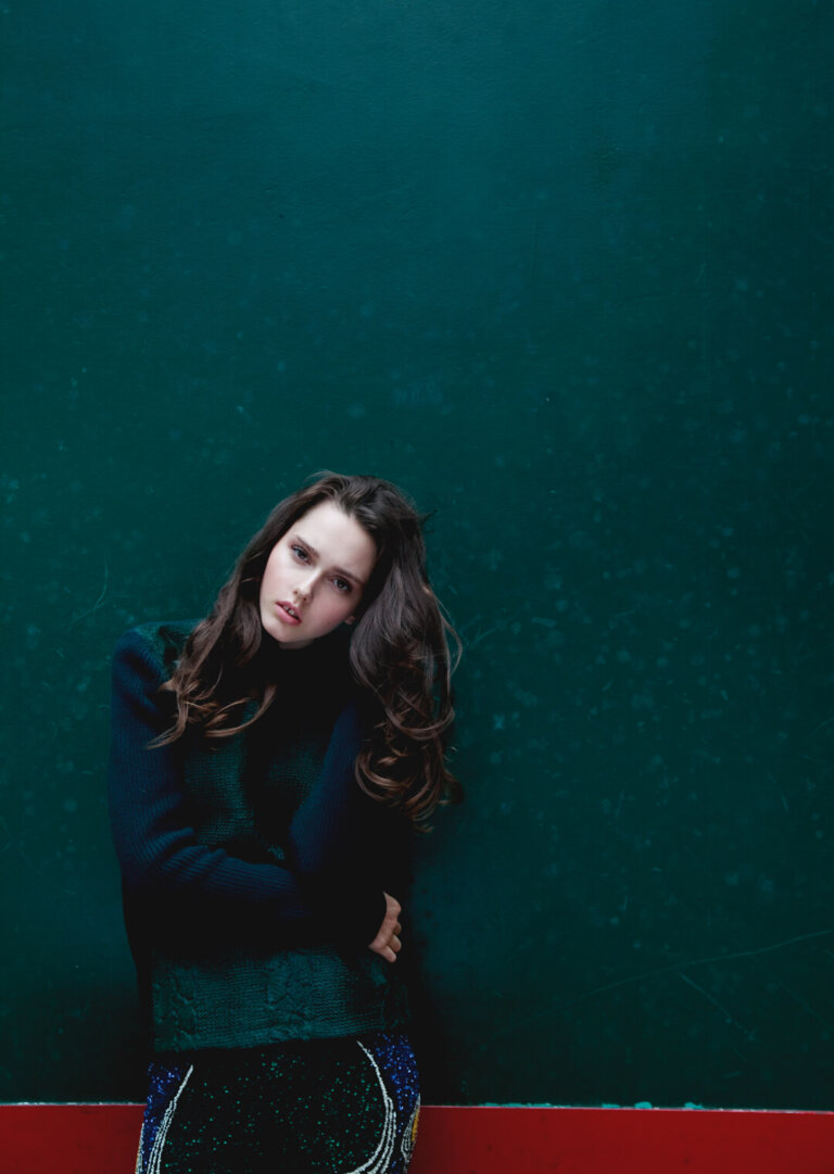 Portrait of a brunette woman in a dark green sweater against a teal background with red stripe at the bottom. Fashion editorial shot.