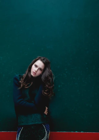 Portrait of a brunette woman in a dark green sweater against a teal background with red stripe at the bottom. Fashion editorial shot.