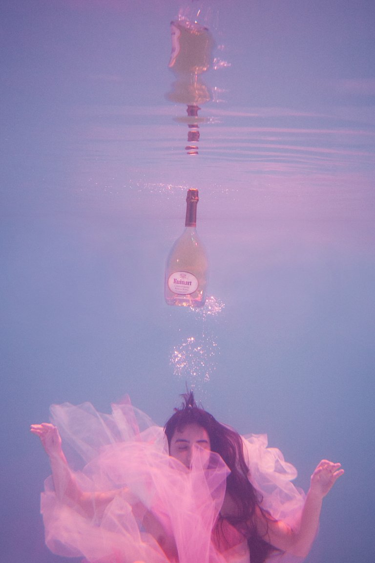 Underwater view of a person in a pink dress, with a floating wine bottle above, creating a surreal, ethereal composition.