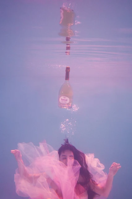 Underwater view of a person in a pink dress, with a floating wine bottle above, creating a surreal, ethereal composition.