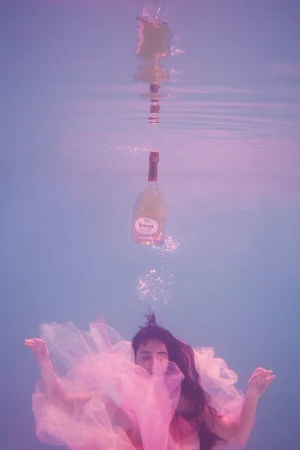 Underwater view of a person in a pink dress, with a floating wine bottle above, creating a surreal, ethereal composition.