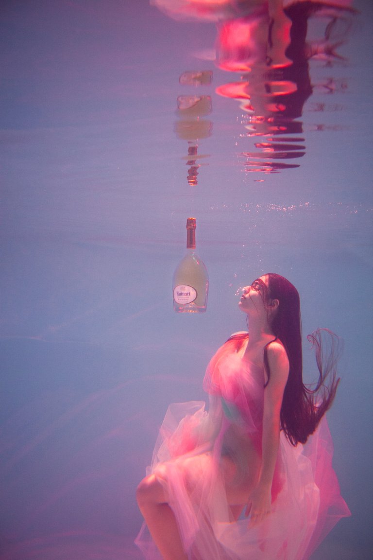 Woman in flowing pink dress floating underwater beneath a suspended glass bottle by Nastasia Dusapin
