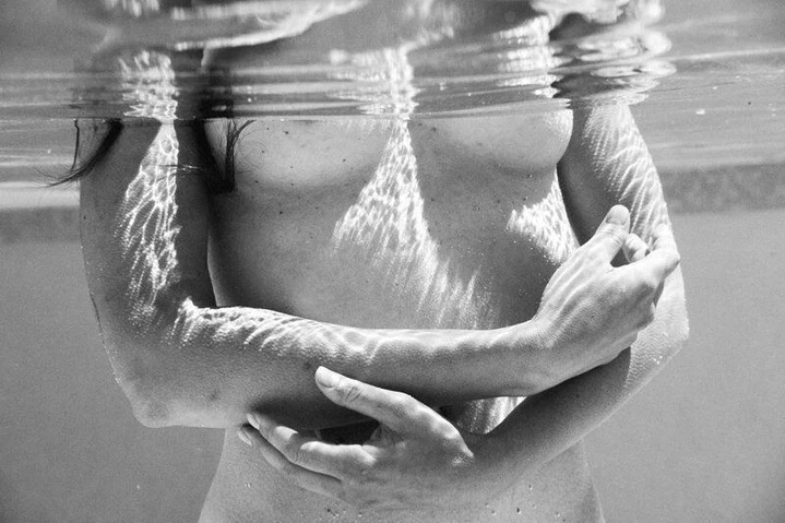 Monochrome photo of a person underwater, arms crossed, with light patterns playing across skin and the surface of the water above.