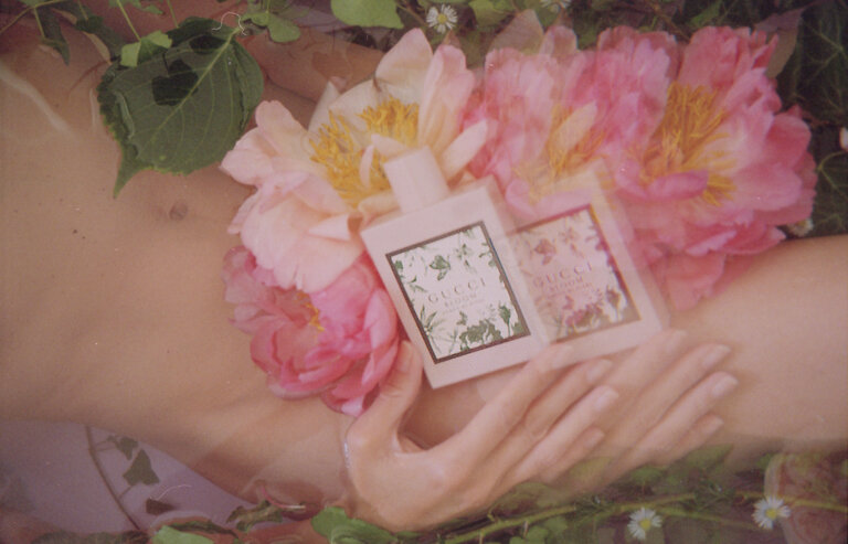 Overhead shot: two perfume bottles surrounded by pink flowers and lush green foliage, nestled in an unidentifiable body of water.