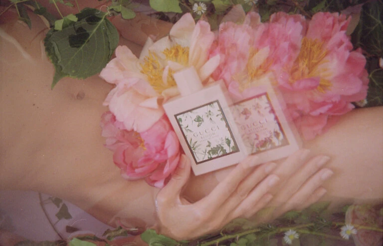 Overhead shot: two perfume bottles surrounded by pink flowers and lush green foliage, nestled in an unidentifiable body of water.