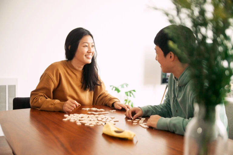 Two smiling people play a game with letter tiles on a wood table in a brightly lit interior. A vase with plants is in the foreground.