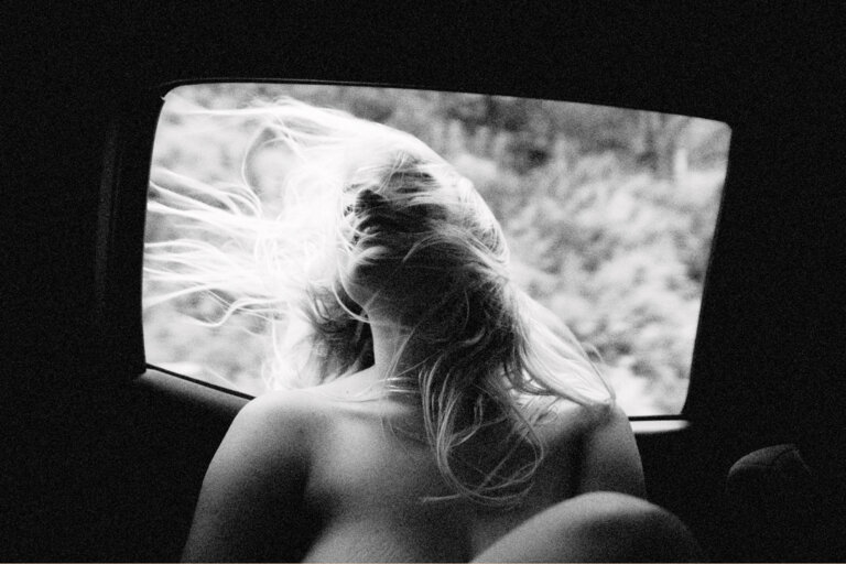 Black and white image of a woman with long hair blowing in the wind, viewed from inside a car, with a blurred nature scene visible.