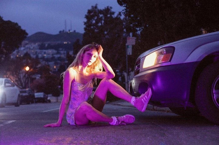 A young adult woman in a dress and sneakers sits on an asphalt road near a car under dark lighting in an urban environment at dusk.