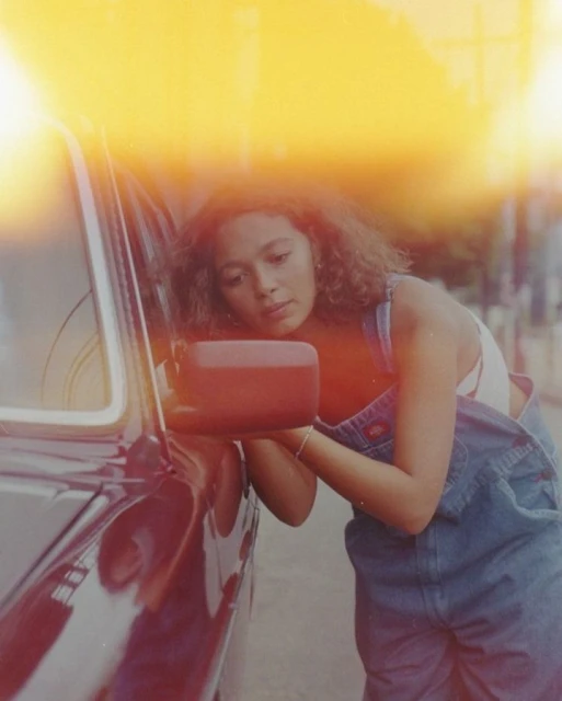 Young woman with curly hair looks in the side mirror of a maroon car outdoors; she wears denim overalls with a white tank top.