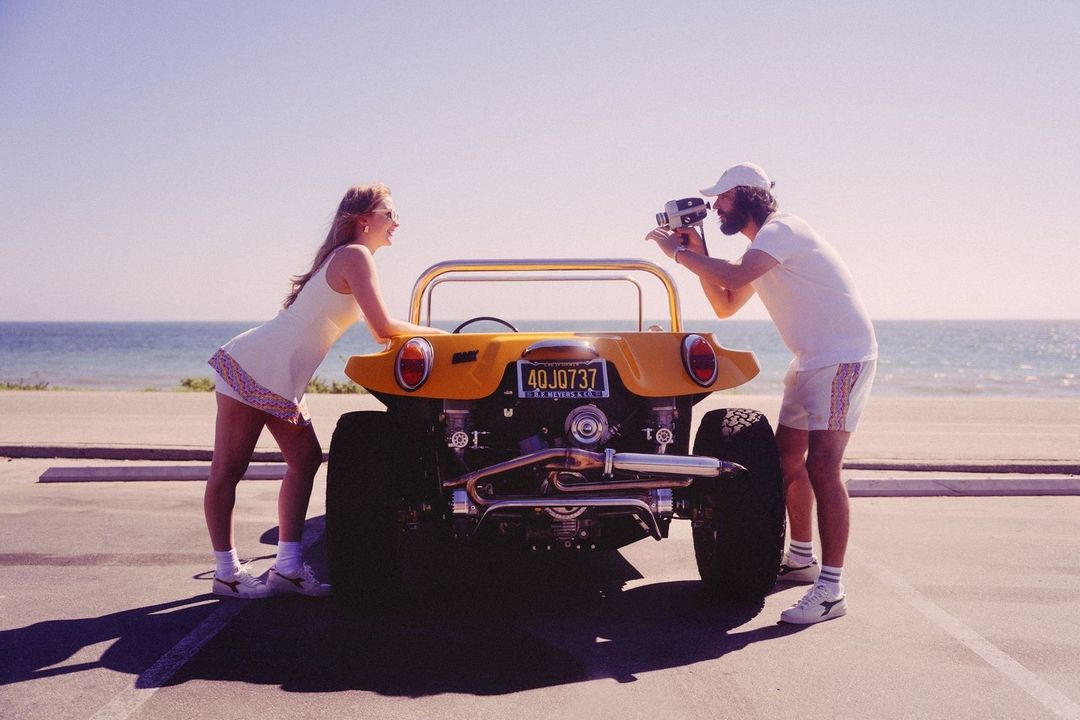 A blonde woman leans against an open-topped yellow dune buggy, while a bearded man films with a vintage camera, on a sunny beach.