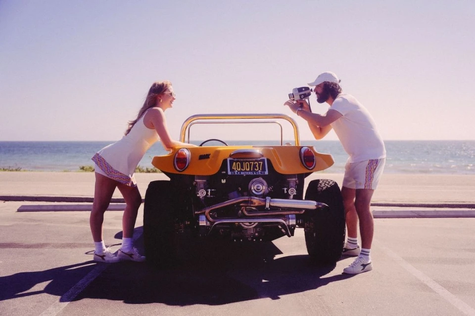 A blonde woman leans against an open-topped yellow dune buggy, while a bearded man films with a vintage camera, on a sunny beach.