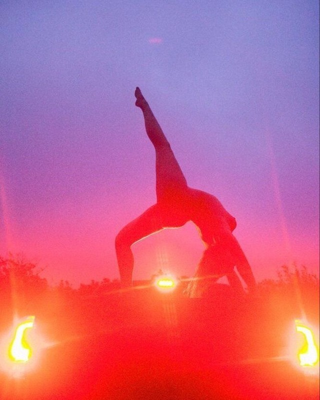 Silhouette of an adult woman performing a yoga pose on a vehicle at dusk, with a pink and purple sky and illuminated lights.