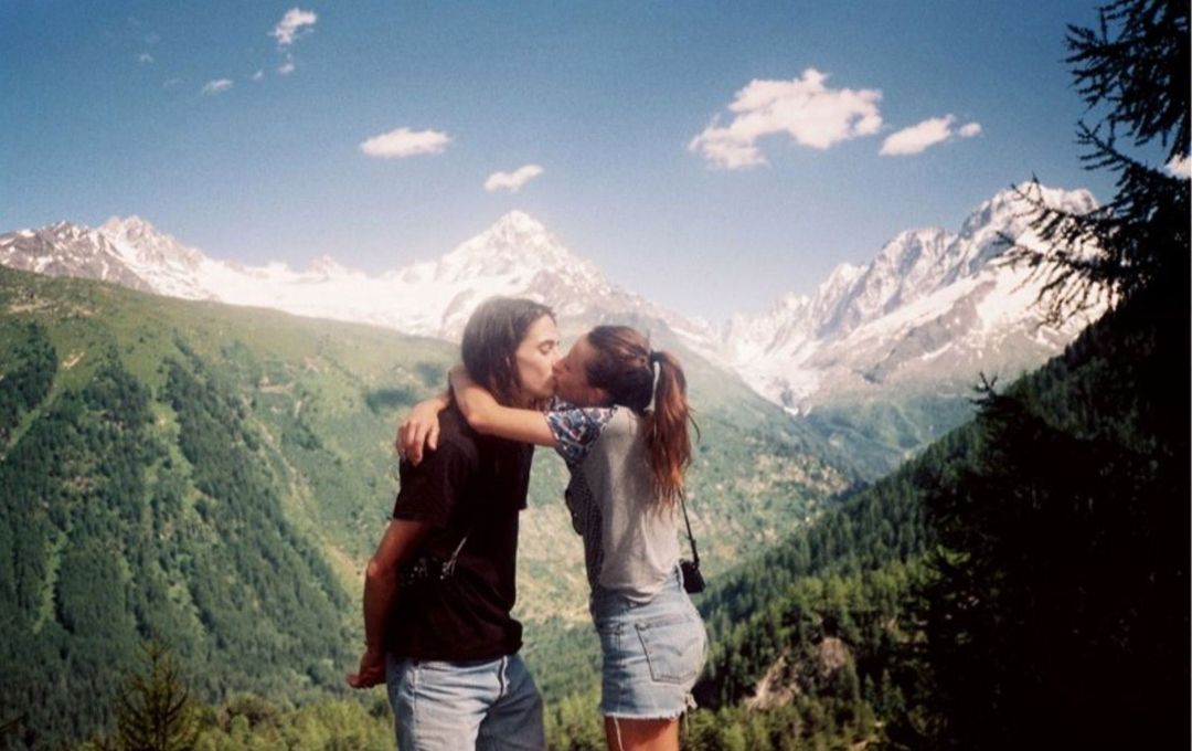 A couple embraces and kisses in a mountainous alpine environment under a clear blue sky with snow-capped peaks.