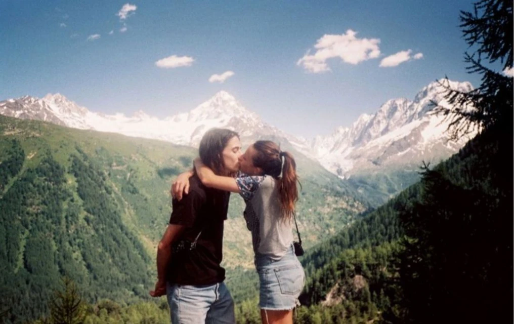 A couple embraces and kisses in a mountainous alpine environment under a clear blue sky with snow-capped peaks.