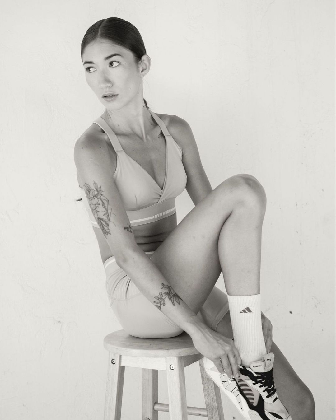 Woman in athleisure wear sits on a wooden stool adjusting an athletic shoe, set against a clean white background in a studio setting.