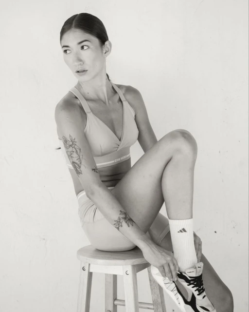 Woman in athleisure wear sits on a wooden stool adjusting an athletic shoe, set against a clean white background in a studio setting.