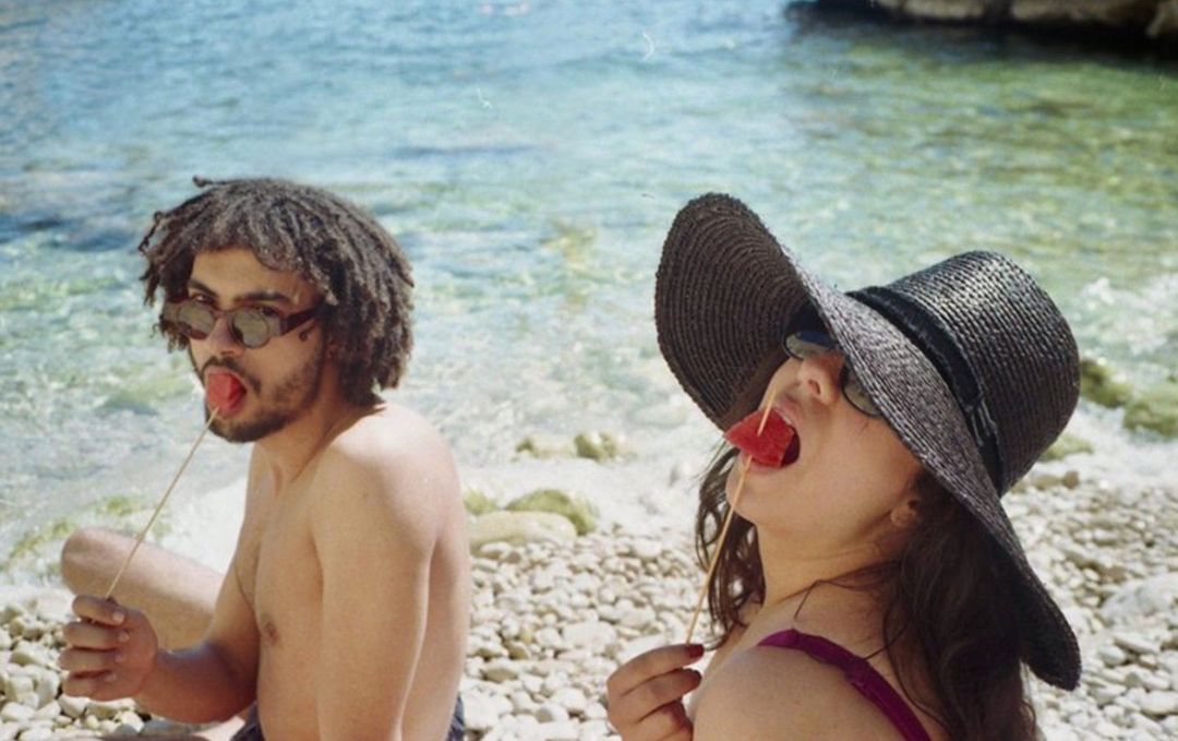 Two people, an adult male and an adult female, eat watermelon on a rocky beach with a sunny ocean view and a clear sky background.
