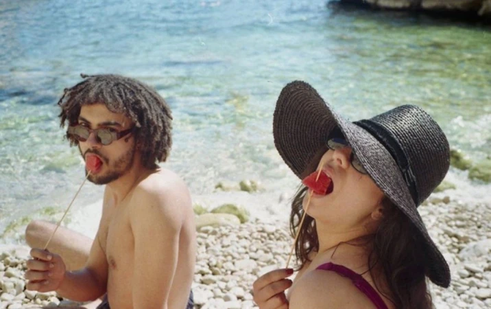 Two people, an adult male and an adult female, eat watermelon on a rocky beach with a sunny ocean view and a clear sky background.