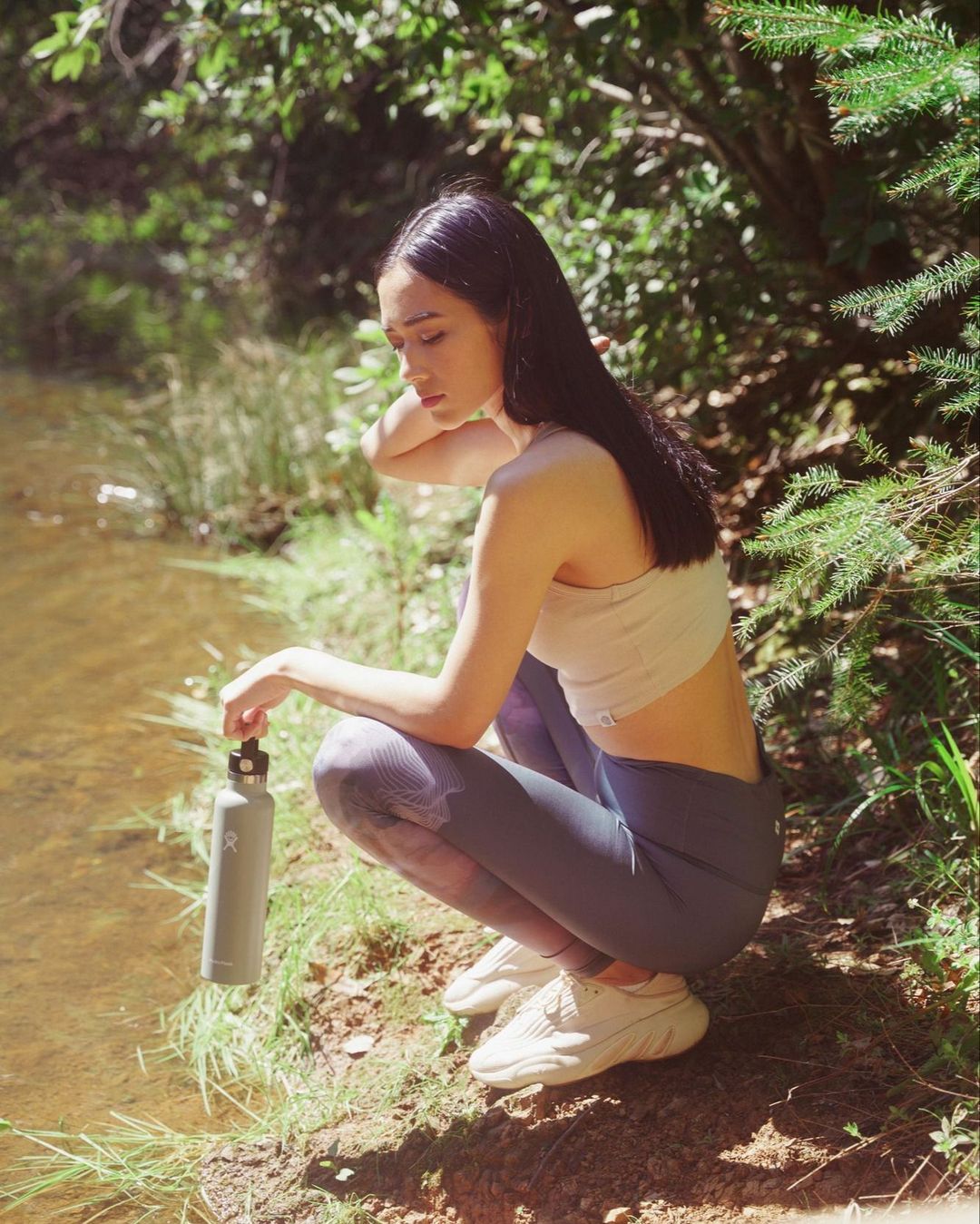 Woman in activewear crouching by a riverbank holding a water bottle amidst trees.