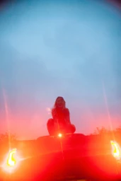 A woman crouching on top of a car roof against a colorful sunset sky with strong orange light flare.