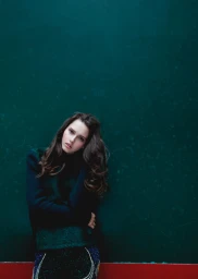 Woman with long dark hair wearing a dark green top posing against a dark teal wall photographed by Nastasia Dusapin.