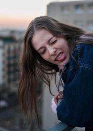 Young woman in a denim jacket sticking out her tongue outdoors against a blurred cityscape background.