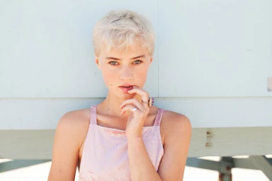 Portrait of a young woman with short blonde hair wearing a pink top and rings against a light wall