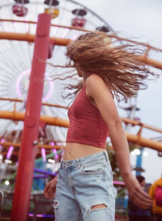 Woman with long wavy hair whipping her head around wearing a red crop top and ripped denim jeans with a Ferris wheel in the background.