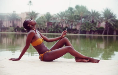 Black woman wearing a yellow bikini sitting poolside next to a body of water with palm trees in the background