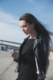 Woman with long brown hair wearing a black leather jacket holding a perfume bottle under a clear sky.