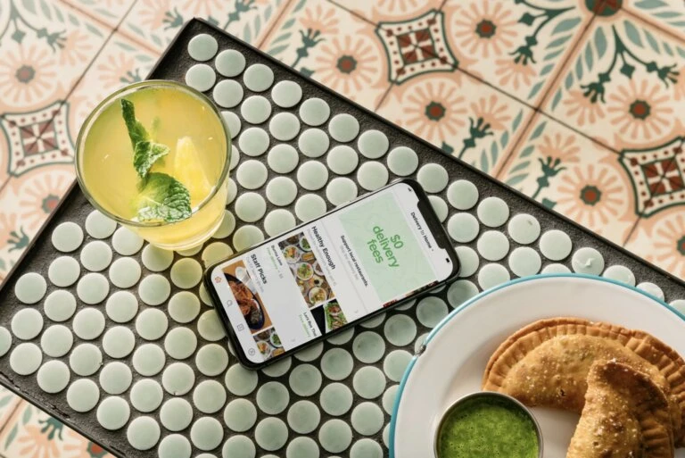 Overhead shot of a smartphone showing a food delivery app, a cocktail, and a plate of empanadas on a colorful patterned table.