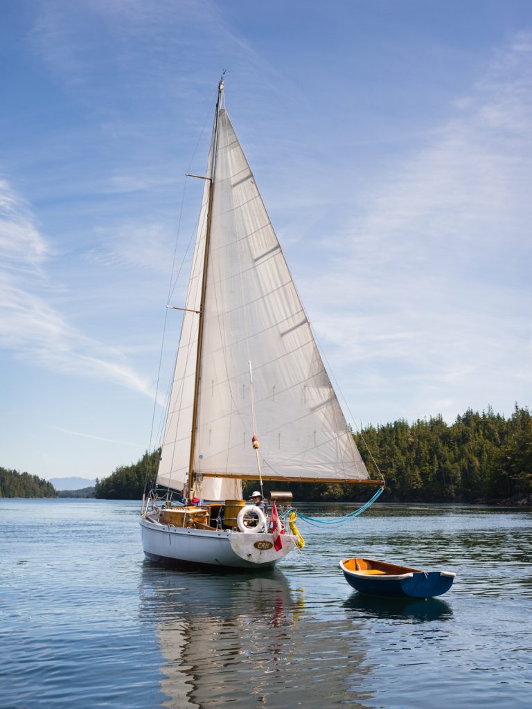 A white sailboat with a tall mast sails on calm blue water near a wooded shoreline under a partly cloudy, sunny sky with a small dinghy.