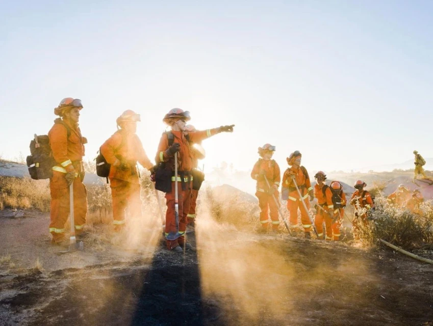 Group of firefighters in orange uniforms standing on a hill, possibly assessing a wildfire situation in natural outdoor setting.