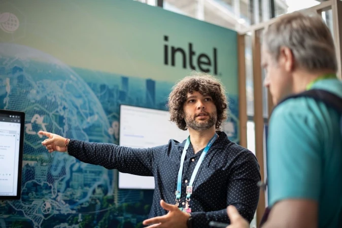 Two men at a tech conference stand in front of a screen and an Intel display, one pointing toward the screen, both in conversation.