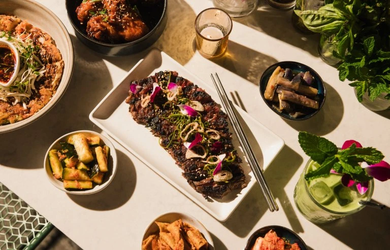 Overhead shot of a curated assortment of Korean dishes, including grilled beef, kimchi, and green matcha cocktails on a table.