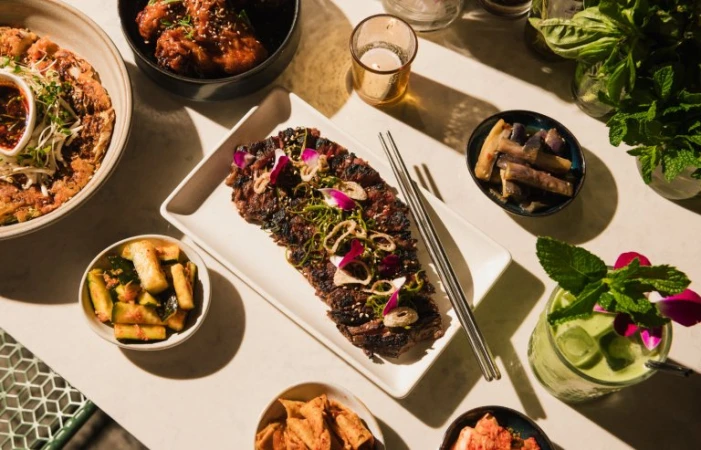 Overhead shot of a curated assortment of Korean dishes, including grilled beef, kimchi, and green matcha cocktails on a table.